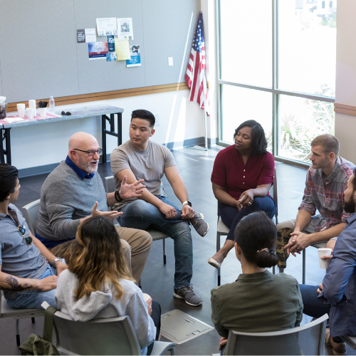 Diverse group of veterans and community members engaged in a discussion in a circle, with the American flag in the background, during an AboutFace-USA® educational program.